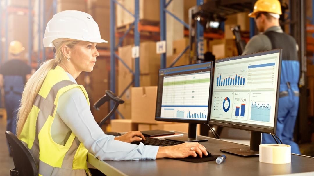 Foto einer Mitarbeiterin im Lager eines Unternehmens am Bildschirm, auf dem statistische Auswertungen einer Anwendung für Logistik und Lagerhaltung zu sehen sind