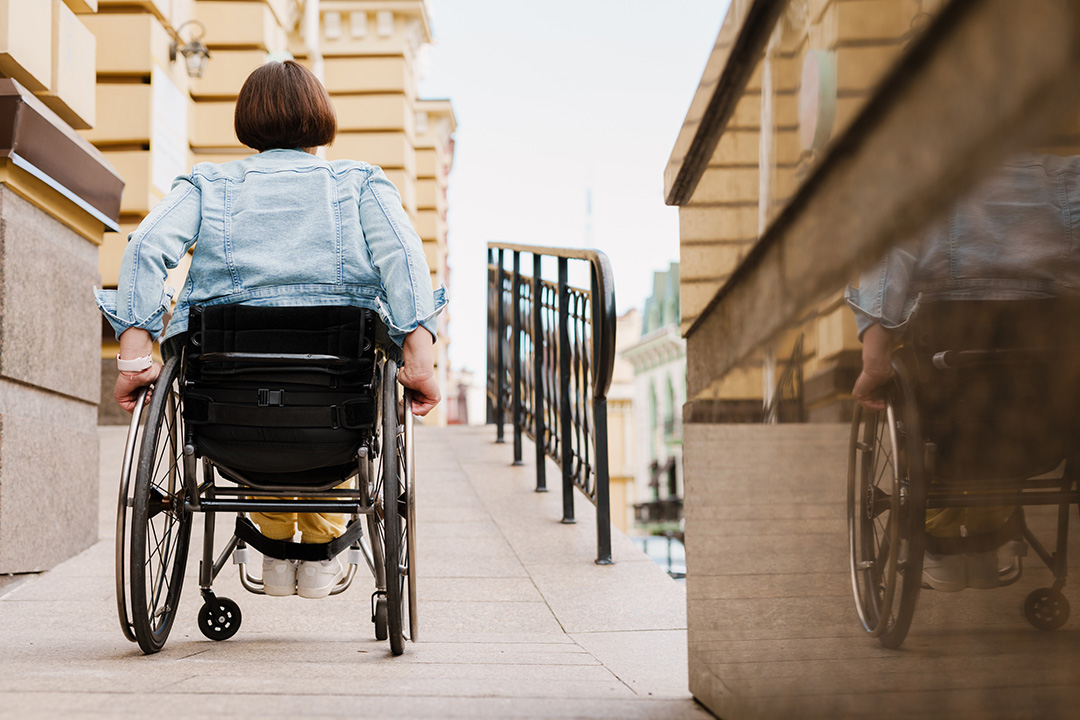 Foto einer Rollstuhlfahrerin, die eine langgestreckte gepflasterte Rampe zum Eingang eines Gebäudes hochfährt.